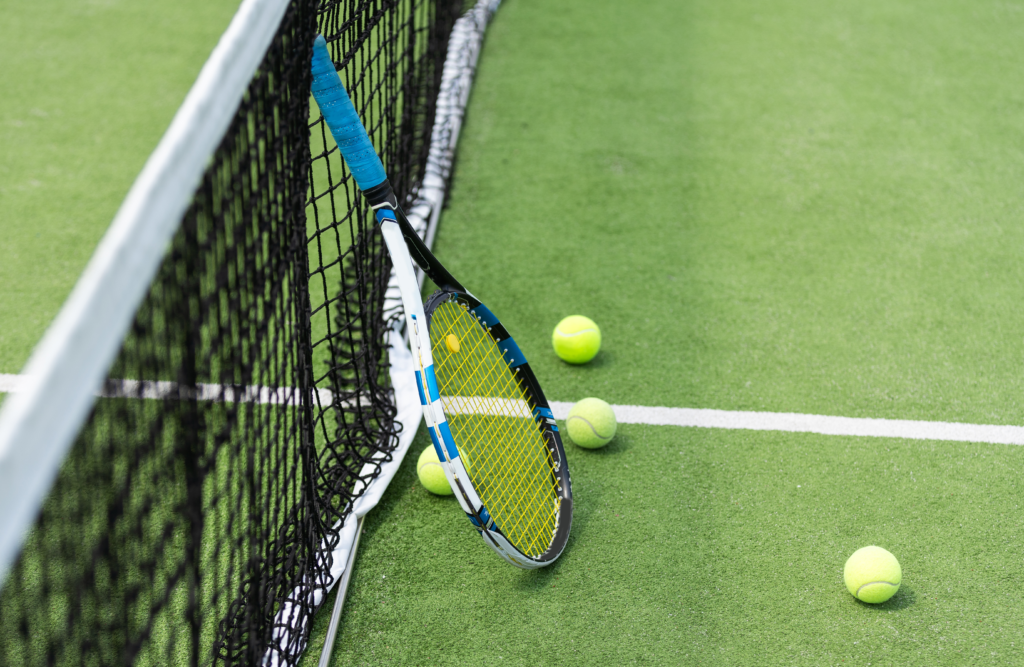 ClubU, Tu Club Exclusivo de Ocio y Deporte Cervera del Maestre. Raqueta de tenis blanca con toques cian apoyada sobre la red de una pista de tenis con 3 pelotas a su alrededor.